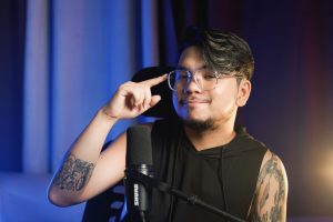 Young man with tattoos and eyeglasses sitting indoors, speaking into a microphone. — The Founding of YouTube A Short History