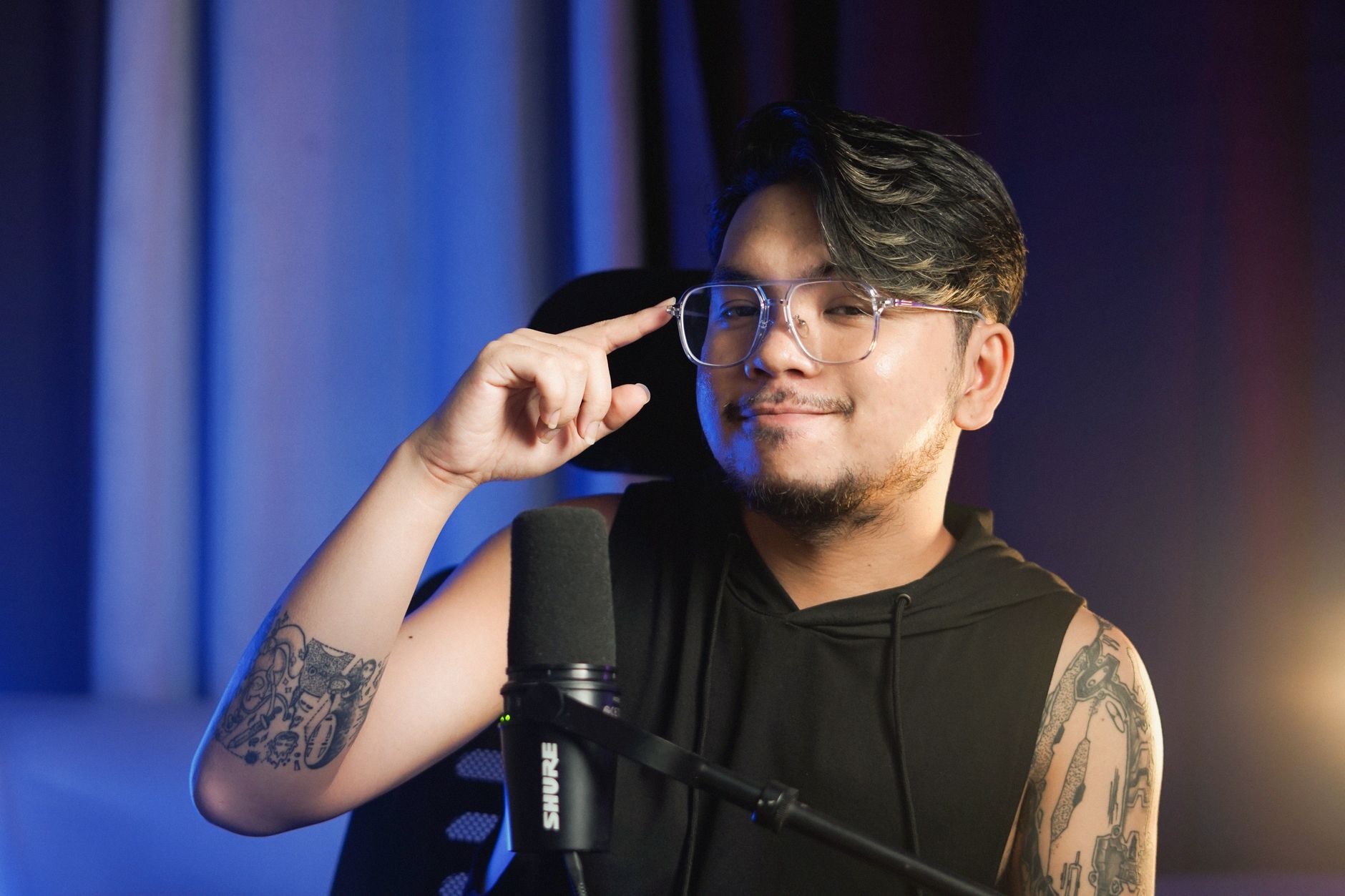 Young man with tattoos and eyeglasses sitting indoors, speaking into a microphone. — The Founding of YouTube A Short History
