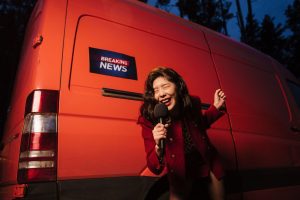 Asian woman reporter energetically broadcasting with a microphone in front of a news van at night. — Wie organisiert Polestar